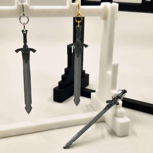 May include: Grey sword-shaped earrings with detailed hilts and blades, displayed on a white and black stand. One pair has silver hooks, the other has gold hooks. A single sword charm rests on the surface.