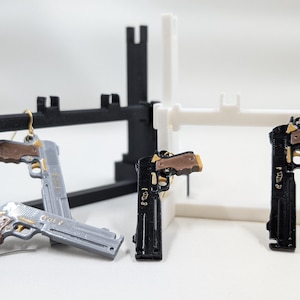May include: Three silver, black and brown gun-shaped earrings hanging from a white and black display stand. The earrings are detailed with gold accents and the text "GOLD" is visible on one of the earrings.
