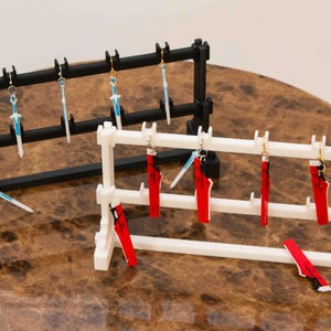 May include: Two earring display stands, one black and one white, with various earrings hanging from them. The earrings are made of metal and have different designs, including swords and red and white patterns.