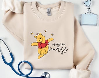 May include: A beige sweatshirt with a Winnie the Pooh character wearing a nurse's hat and a red shirt. The text "PEDIATRIC nurse" is printed on the sweatshirt.