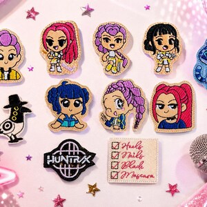 May include: A collection of embroidered patches featuring seven stylized anime-style characters, a blue cat-like creature, a black and white bird, a black logo patch with the text HUNTRIX, and a square patch with a checklist reading Heals, Nails, Blade, Mascara.
