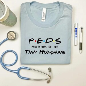 May include: Light blue t-shirt with the text "P.E.D.S. Protectors of the Tiny Humans" in black. The letters "P.E.D.S." are in a stylized font with colored dots.