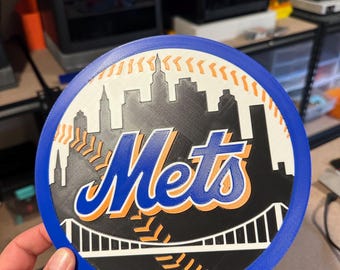 May include: A 3D printed baseball-themed wall decor featuring the New York Mets logo. The logo is in blue, orange, and white with a black background. The design includes a cityscape, a bridge, and baseball stitching.
