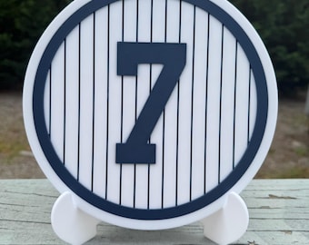 May include: A white and navy blue decorative sign featuring the number 7. The sign has a circular shape with vertical stripes and a navy blue border. It is supported by a white stand.