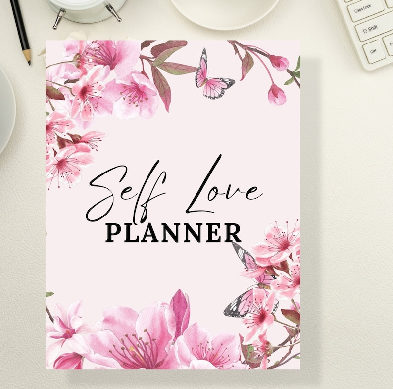 Op de afbeelding: Een roze en witte bloemenplanner met de tekst "Self Love Planner" in zwarte letters. De planner is versierd met vlinders en roze bloemen.
