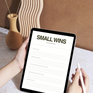 May include: A digital tablet displaying the words "SMALL WINS" in a dark green font. The tablet is held by a person, with a stylus in the other hand. A decorative vase with dried flowers and a wooden wave-shaped decor are in the background.
