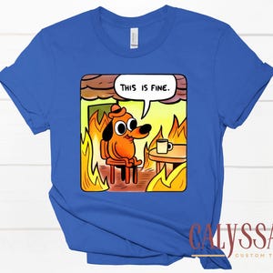 May include: Royal blue t-shirt featuring a cartoon dog in a burning room, with the text "THIS IS FINE" in a speech bubble. The dog is sitting calmly with a coffee cup on a table. The shirt has a crew neck and short sleeves.