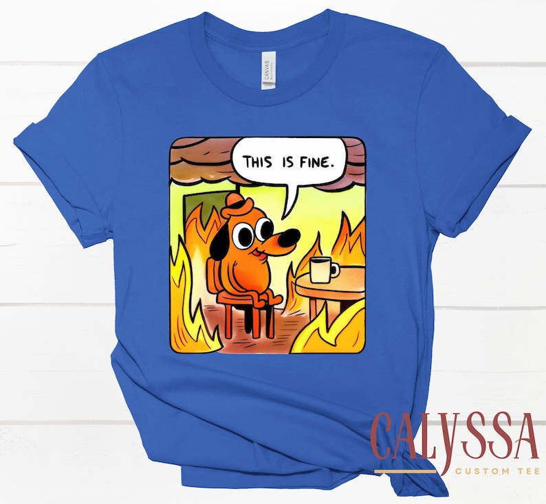 May include: Royal blue t-shirt featuring a cartoon dog in a burning room, with the text "THIS IS FINE" in a speech bubble. The dog is sitting calmly with a coffee cup on a table. The shirt has a crew neck and short sleeves.