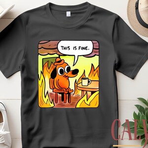 May include: Black t-shirt featuring a cartoon dog in a burning room, with the text "THIS IS FINE" in a speech bubble. The dog is orange and wearing a hat, sitting in a chair with a coffee cup on a table. The shirt is a custom tee.