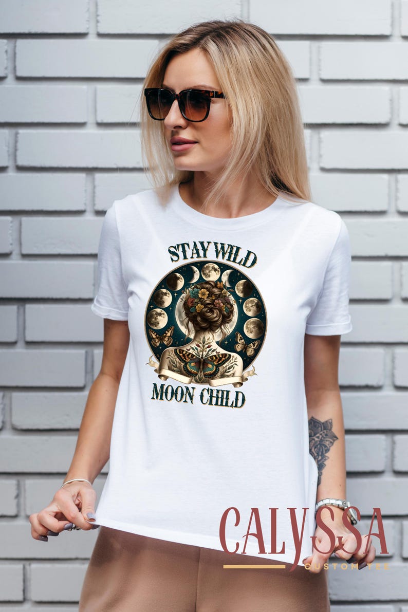 May include: White t-shirt with a graphic print of a woman with flowers in her hair and butterfly tattoos on her back. The graphic is surrounded by a circle of moons. The text on the shirt reads 'Stay Wild Moon Child'.