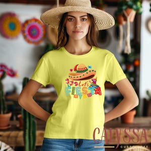 May include: A yellow t-shirt featuring a sombrero and the words "Let's Fiesta" in colorful letters. The shirt has a crew neck and short sleeves. The design includes maracas, a pinata, and other festive elements.