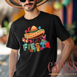 May include: Black t-shirt with a colorful "Let's Fiesta" graphic. The design includes a sombrero, maracas, and a pinata. The shirt is a crew neck style and is made of a soft material.
