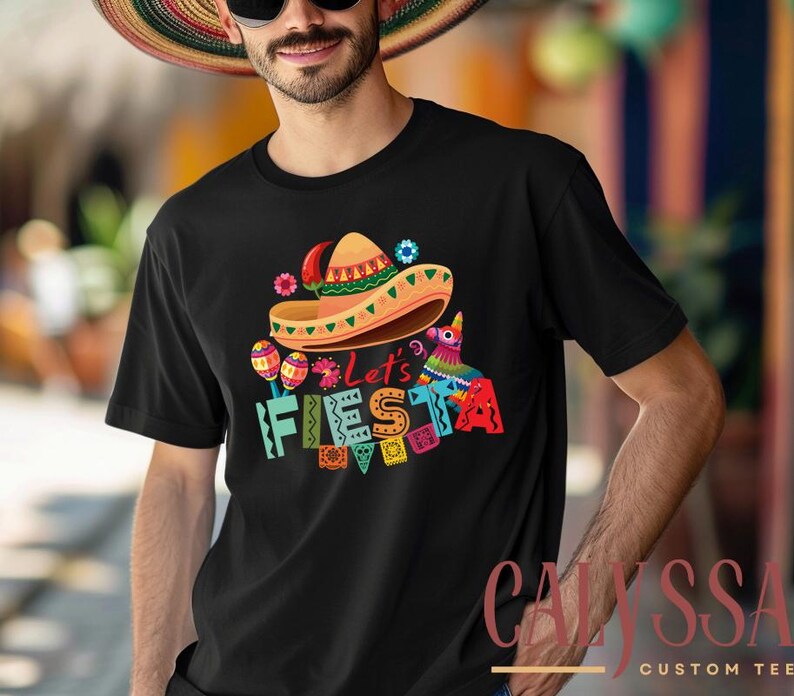 May include: Black t-shirt with a colorful "Let's Fiesta" graphic. The design includes a sombrero, maracas, and a pinata. The shirt is a crew neck style and is made of a soft material.
