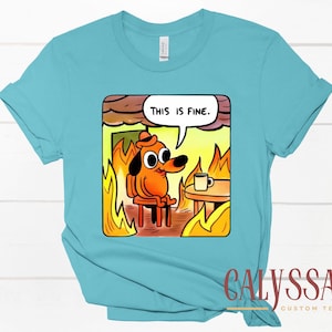 May include: A light blue t-shirt featuring a cartoon dog sitting in a burning room, with the text "THIS IS FINE" in a speech bubble. The dog is orange and wearing a hat, with a coffee cup on a table nearby.