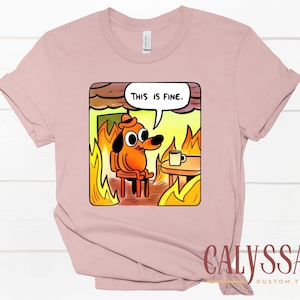 May include: A light pink t-shirt featuring a cartoon dog sitting in a burning room, with the text "THIS IS FINE" in a speech bubble. The dog is wearing a hat and there is a coffee cup on a table.