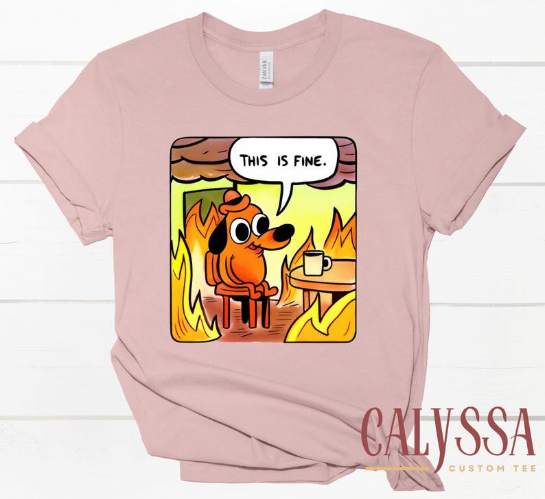 May include: A light pink t-shirt featuring a cartoon dog sitting in a burning room, with the text "THIS IS FINE" in a speech bubble. The dog is wearing a hat and there is a coffee cup on a table.