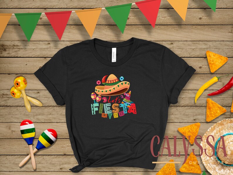 May include: Black t-shirt with a colorful "Let's Fiesta" graphic featuring a sombrero, maracas, and a pi&ntilde;ata. The shirt is decorated with festive elements, perfect for a celebration.