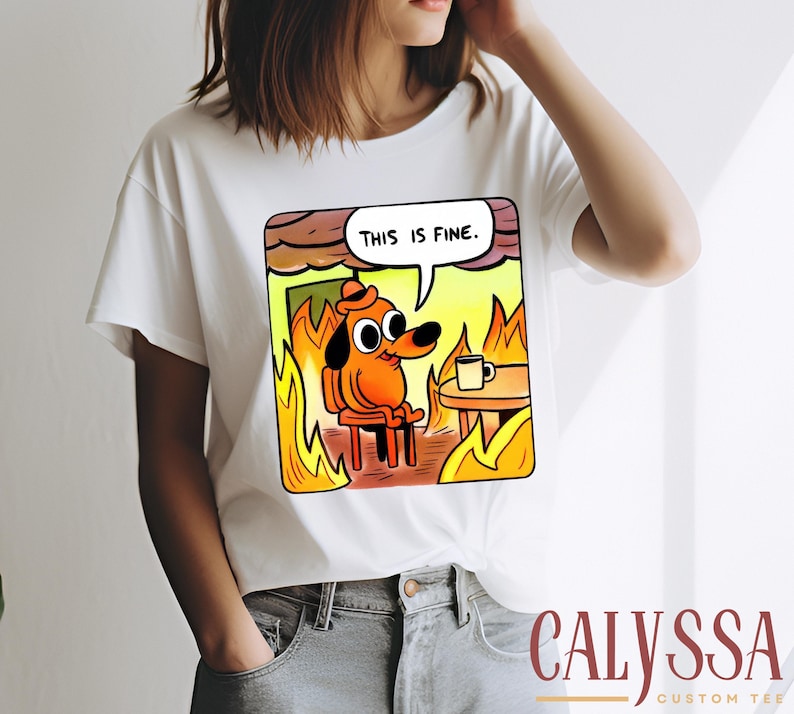 May include: White t-shirt featuring a cartoon dog in a burning room, with the text "THIS IS FINE" in a speech bubble. The dog is sitting in a chair with a coffee cup on a table. The shirt has the text "CALYSSA CUSTOM TEE" at the bottom.