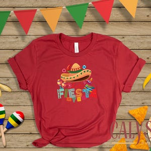 May include: Red t-shirt with a colorful "Fiesta" graphic featuring a sombrero, maracas, and other festive elements. The image includes tortilla chips, chili peppers, and a decorative banner, evoking a celebratory theme.