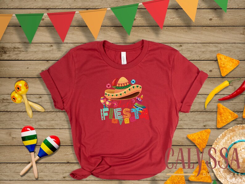 May include: Red t-shirt with a colorful "Fiesta" graphic featuring a sombrero, maracas, and other festive elements. The image includes tortilla chips, chili peppers, and a decorative banner, evoking a celebratory theme.