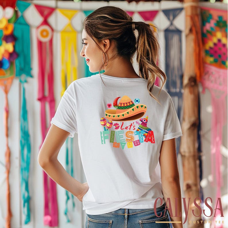 May include: White t-shirt with a colorful graphic on the back featuring a sombrero, maracas, and the words "Let's Fiesta". The design includes a pi&ntilde;ata and festive banner elements, perfect for a celebration.