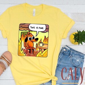 May include: Yellow t-shirt featuring a cartoon dog in a burning room, with the text "THIS IS FINE" in a speech bubble. The shirt is paired with blue denim shorts and sandals.