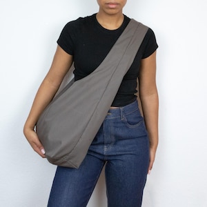 May include: A person wearing a black t-shirt and blue jeans is carrying a large, gray, fabric tote bag over their shoulder.