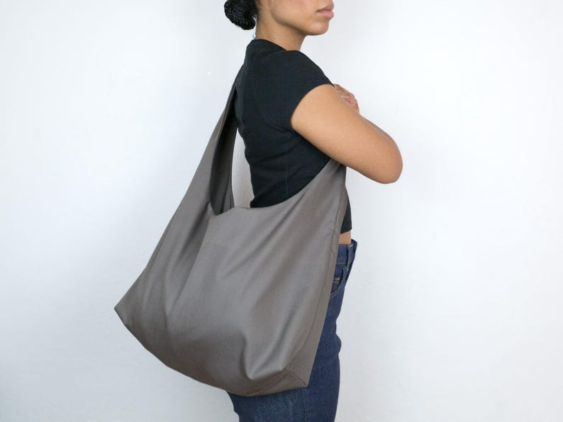 May include: A large, gray, fabric tote bag with a wide opening and a single strap. The bag is worn over the shoulder of a person wearing a black shirt and blue jeans.