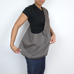 May include: A person wearing a black t-shirt and gray jeans is holding a gray canvas tote bag.