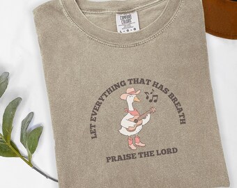 May include: A tan Comfort Colors t-shirt featuring a cartoon goose playing a guitar, wearing a cowboy hat and boots. The text reads "Let everything that has breath praise the Lord."