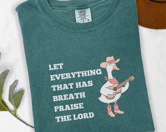 May include: Blue spruce Comfort Colors t-shirt with the text "LET EVERYTHING THAT HAS BREATH PRAISE THE LORD." A cartoon goose wearing a cowboy hat, scarf, and boots plays a ukulele. A brown hat is in the background.
