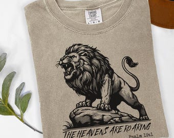 May include: A tan Comfort Colors t-shirt featuring a black illustration of a roaring lion atop a rock. The text "THE HEAVENS ARE ROARING" and "Psalm 19:1" are printed below the lion. The shirt is a size L-G-G.