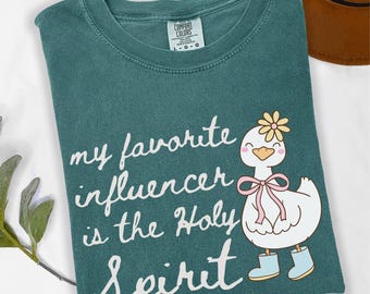 May include: Blue spruce Comfort Colors t-shirt with the text "my favorite influencer is the Holy Spirit." A cartoon duck wearing a flower, bow, and boots is on the right. A brown hat is in the background.