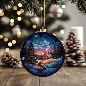 Puede incluir: Adorno navideño redondo con una escena de cabaña invernal. El adorno presenta una cabaña con humo saliendo de la chimenea, rodeada de árboles y un cielo estrellado. El adorno cuelga de una cuerda dorada.