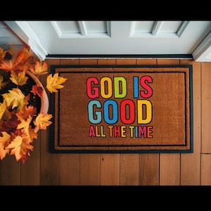 Puede incluir: Una alfombra rectangular marrón con las palabras "GOD IS GOOD ALL THE TIME" en letras de colores. La alfombra tiene un borde negro y está colocada en un suelo de madera cerca de una puerta. Se ve una maceta con hojas de otoño.
