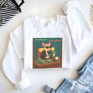 Puede incluir: Sudadera blanca con una imagen de dibujos animados de un personaje con gafas de sol y sosteniendo dinero. La imagen incluye el texto "I worship you, YOU KNOW?" El personaje está sentado en un escritorio con montones de dinero y una taza de café.