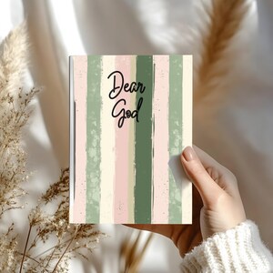 Puede incluir: Un cuaderno con un diseño de rayas verticales en tonos verdes, rosas y crema. Las palabras "Dear God" están escritas en escritura negra en la portada. El cuaderno está en una mano.
