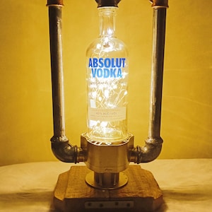 May include: A clear glass bottle with a label that reads "ABSOLUT VODKA" is suspended between two metal pipes. The bottle is illuminated with white fairy lights. The pipes are attached to a wooden base with a metal cap.