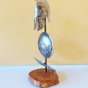 Può includere: Un elmo, uno scudo e una spada spartani color argento sono esposti su una base di legno. L'elmo ha un cimiero e lo scudo presenta un disegno centrale. La spada è posizionata sotto lo scudo.