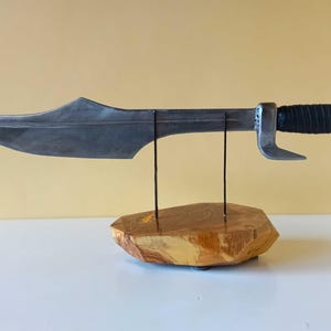 May include: A metal sword with a curved blade and a black wrapped handle. The sword is displayed on a wooden base with two metal supports. The blade has a silver finish.
