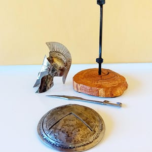 Pode incluir: Um conjunto em miniatura de capacete, escudo e espada espartanos é exibido. O capacete prateado tem uma crista e uma proteção facial. O escudo é redondo com um design em forma de V. Uma espada e uma base de madeira com uma haste preta completam o conjunto.