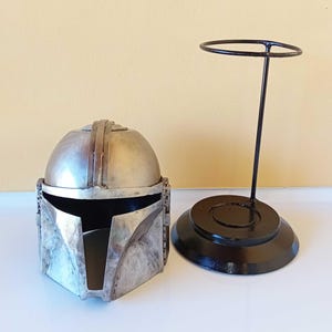 May include: A silver-colored Mandalorian helmet with a matching black display stand. The helmet has a detailed, battle-worn appearance. The stand features a circular base and a vertical support with a ring at the top, designed to hold the helmet.