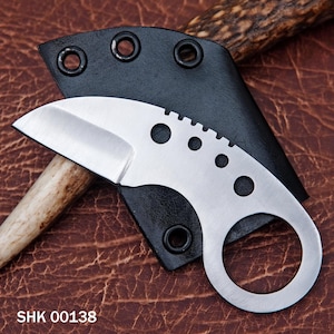Könnte beinhalten: Ein kleines, silbernes, gebogenes Messer mit einer schwarzen Lederscheide. Das Messer hat einen Ring am Griff und eine gezahnte Klinge. Der Text "SHK 00138" ist im Bild sichtbar.