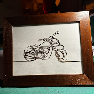 Może przedstawiać: Srebrna rzeźba motocykla z drutu w brązowej drewnianej ramie. Motocykl wykonany jest z srebrnego drutu i utrzymany w minimalistycznym stylu.