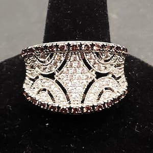 May include: A silver-toned ring with an intricate openwork design. The ring features a band of small, dark red stones along the top and bottom edges. The center of the ring is adorned with clear, sparkling stones, creating a striking contrast.