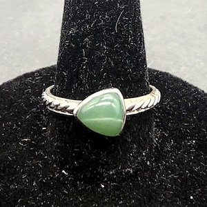 May include: A silver ring with a triangular, light green gemstone. The ring is displayed on a black velvet ring holder. The band has a textured design. The gemstone is smooth and polished.