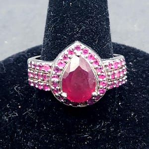 May include: A silver-toned ring featuring a large, pear-shaped, deep red center stone surrounded by a halo of smaller, round, vibrant pink stones. The band is wide and split into three rows, each encrusted with numerous small, round pink stones.