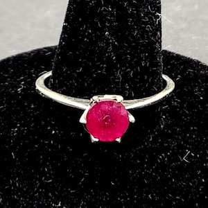 May include: A silver ring featuring a round, deep red gemstone. The ring is presented on a black velvet ring stand. The gemstone's colour is the main focus, with a simple, elegant setting.