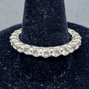 May include: A silver ring with a continuous row of clear gemstones. The ring is displayed on a black velvet ring holder against a dark gray background. The gemstones are round and evenly spaced around the band.