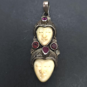 Peut inclure: Pendentif en argent orné de deux visages sculptés aux expressions sereines. Le pendentif est orné de pierres rouges et de motifs métalliques complexes. Les visages sont de couleur ivoire crémeux, sur fond sombre.
